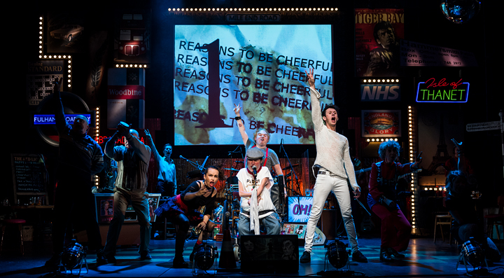All performers on a cluttered and busy stage, with their arms outstretched to the air. The text behind them reads Reasons to be Cheerful.