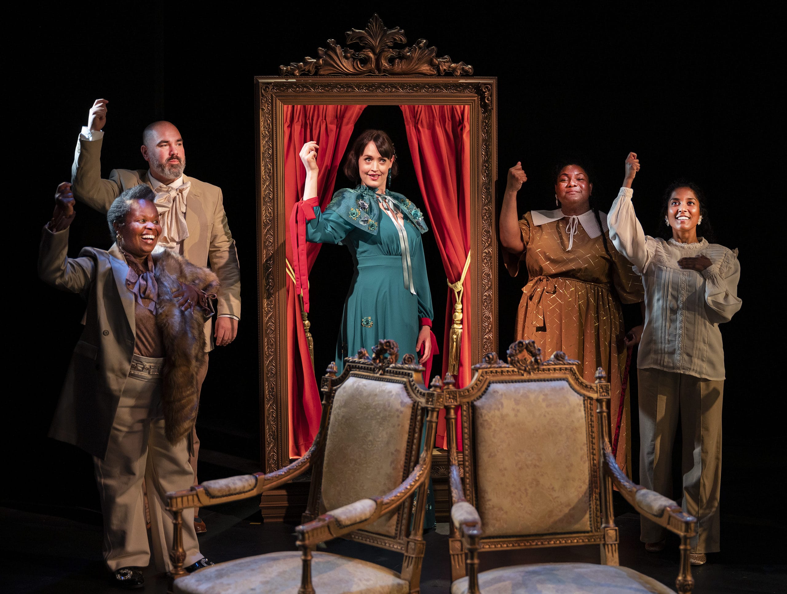 Four performers on stage, they all click their fingers in unison. The performer centre stage emerges from a framed set of curtains, and is wearing a stand-out, turquoise ball gown.