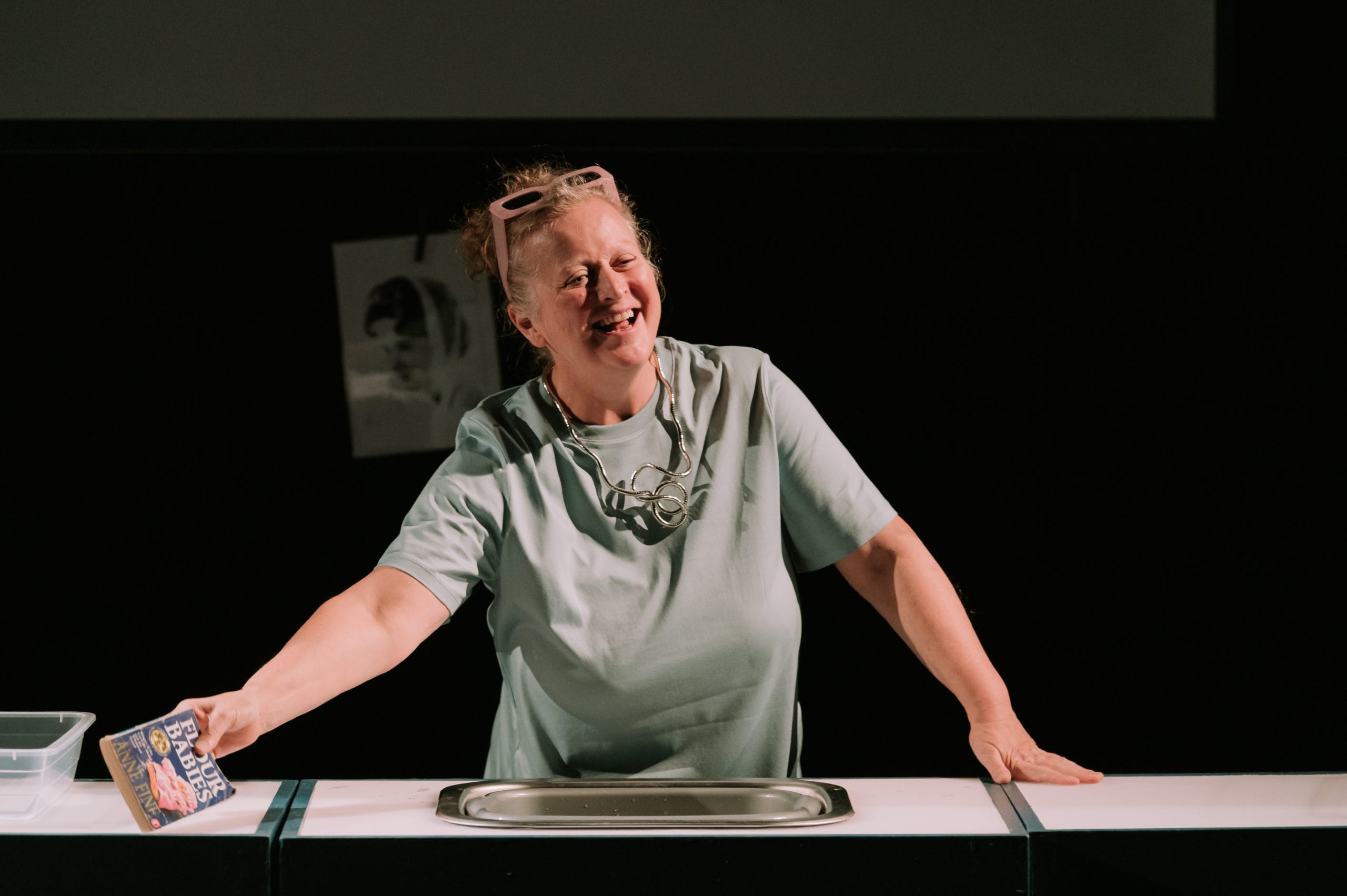 A smiling woman leans over a sink, holding a book in an outstretched arm.