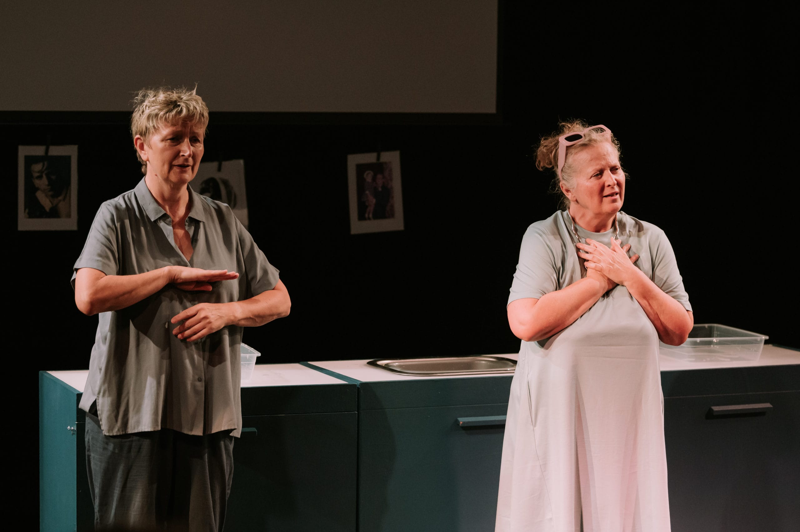 Two women stand on stage; one is signing, the other is clutching her chest.