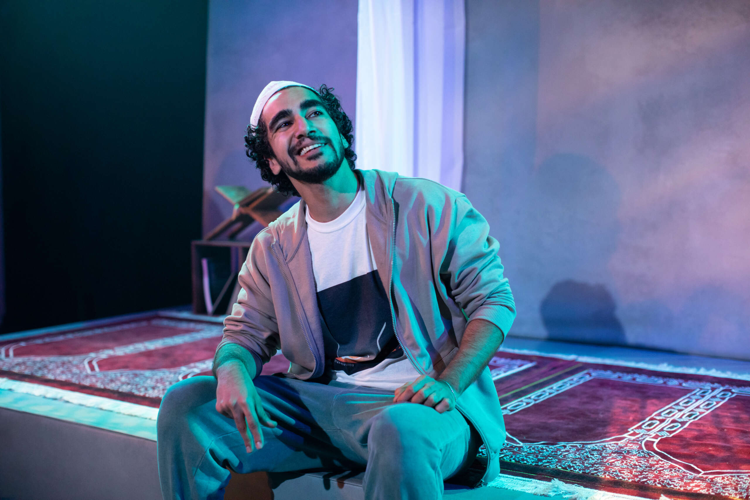 Zaqi Ismail, a young east Asian man with a thick black beard, sits on the edge of the stage. He is wearing a grey matching tracksuit.