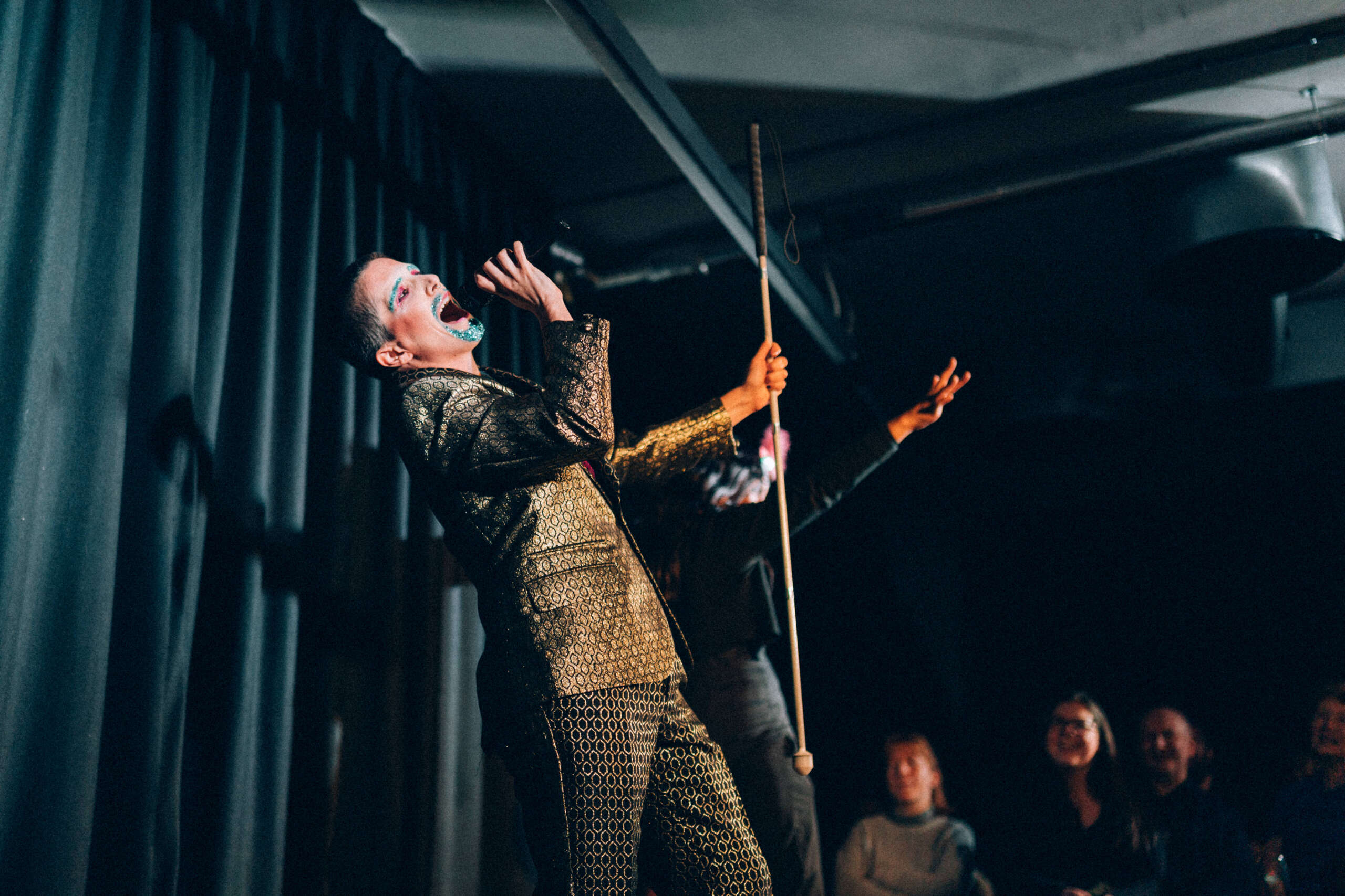 A performer sings passionately into a microphone. They wear a gold sparkly jacket and has blue glittery makeup on their face.