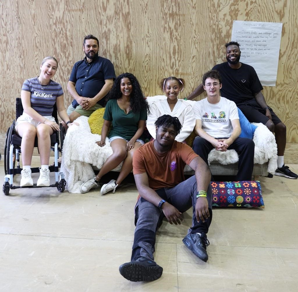 A group of actors and the director sit together smiling to camera