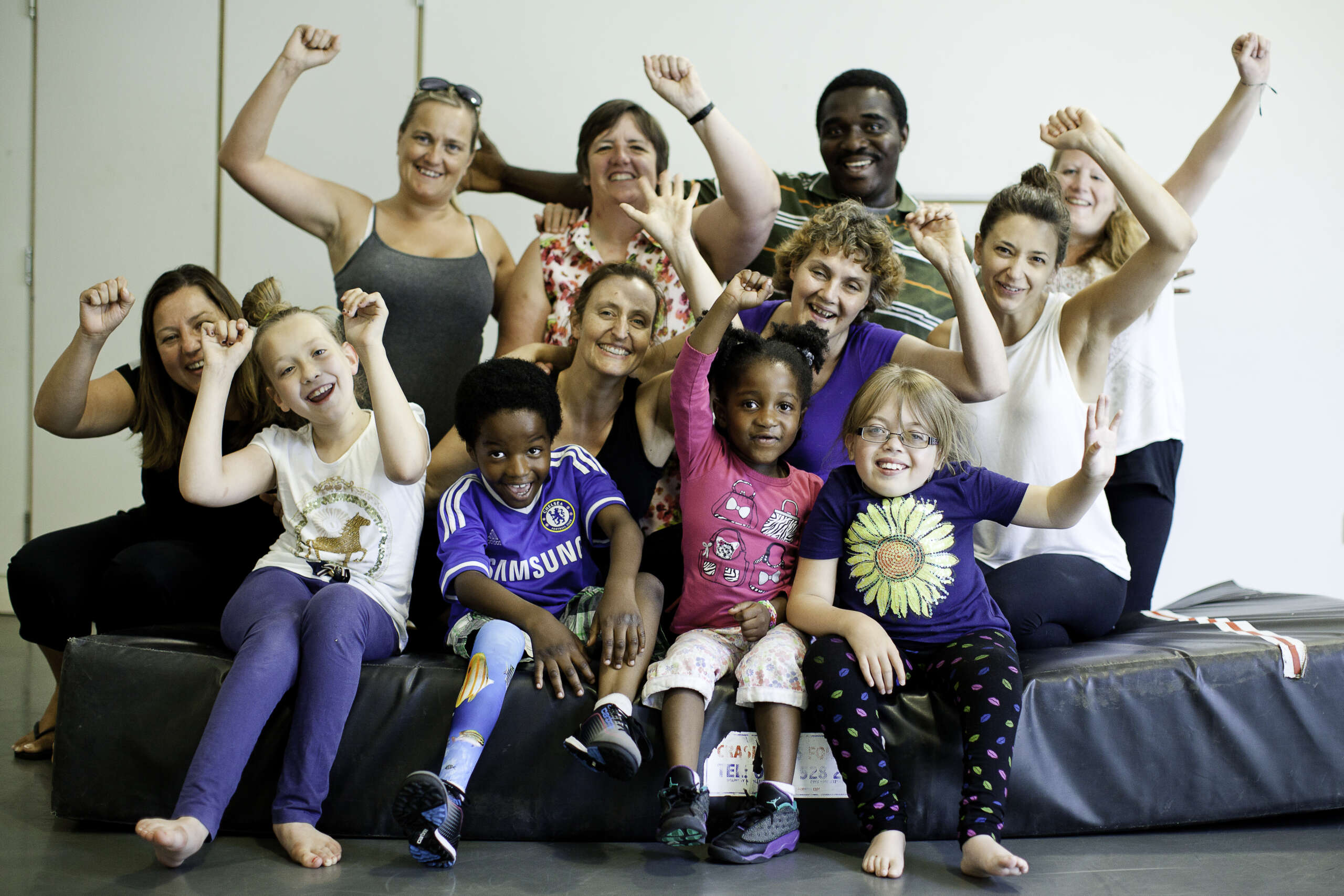 A group of children and adults all sat on a crash mat. They have their fists in the air and are smiling and cheering.