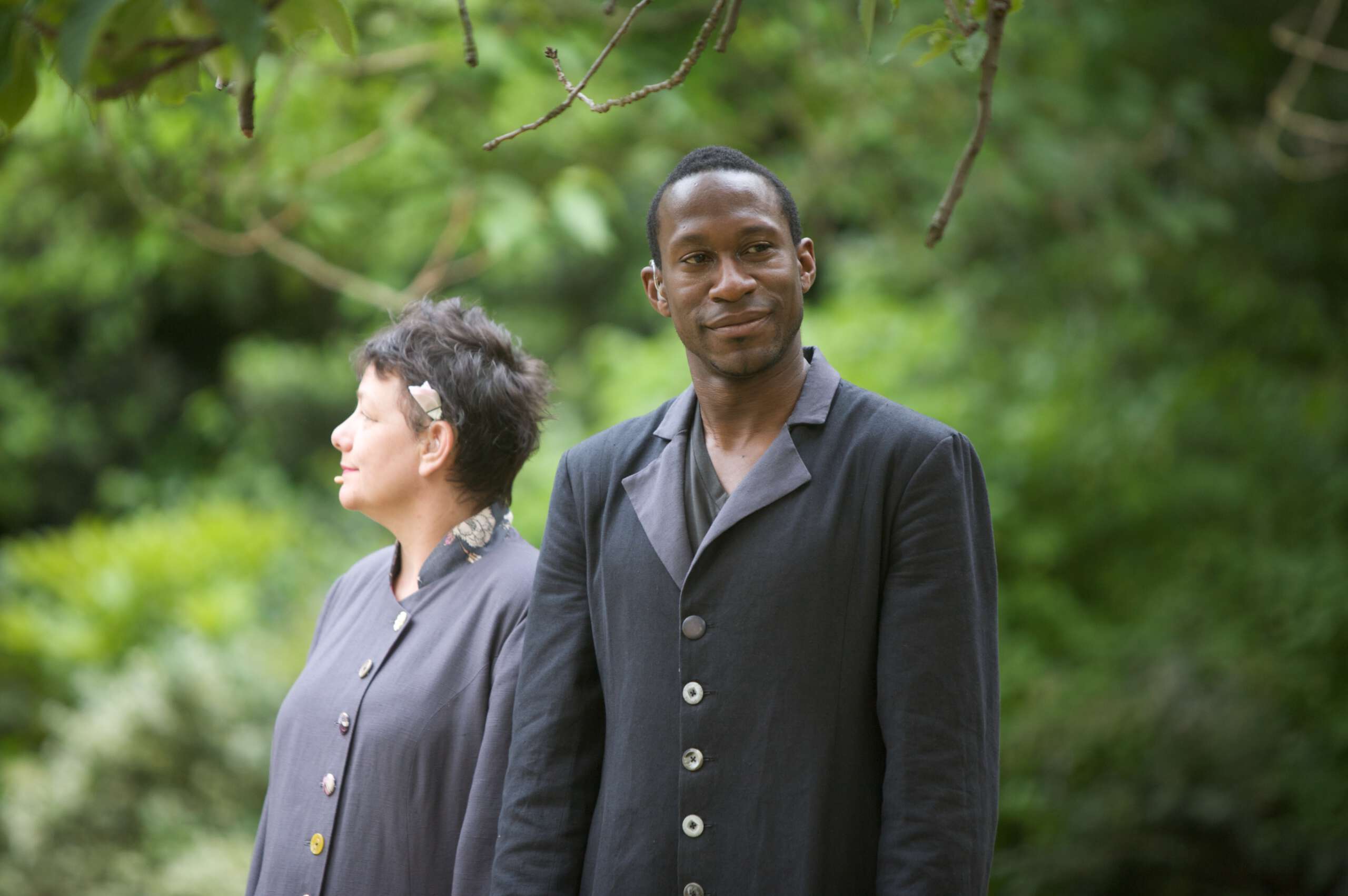 A picture of David Ellington, standing outside with another performer. David is Black, with short hair, and he wears a black suit.