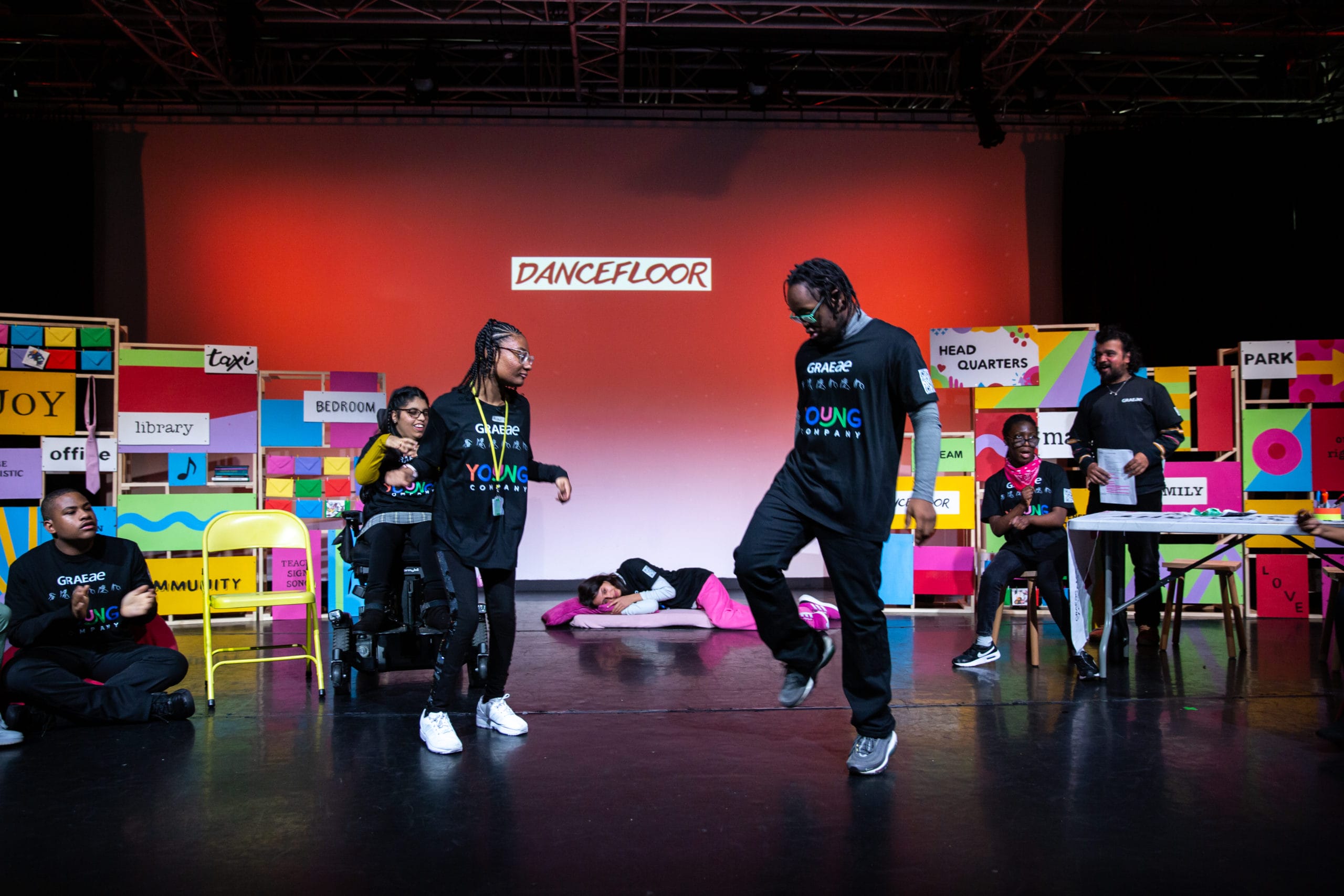 Two performers stand centre stage, smiling at eachother and dancing.