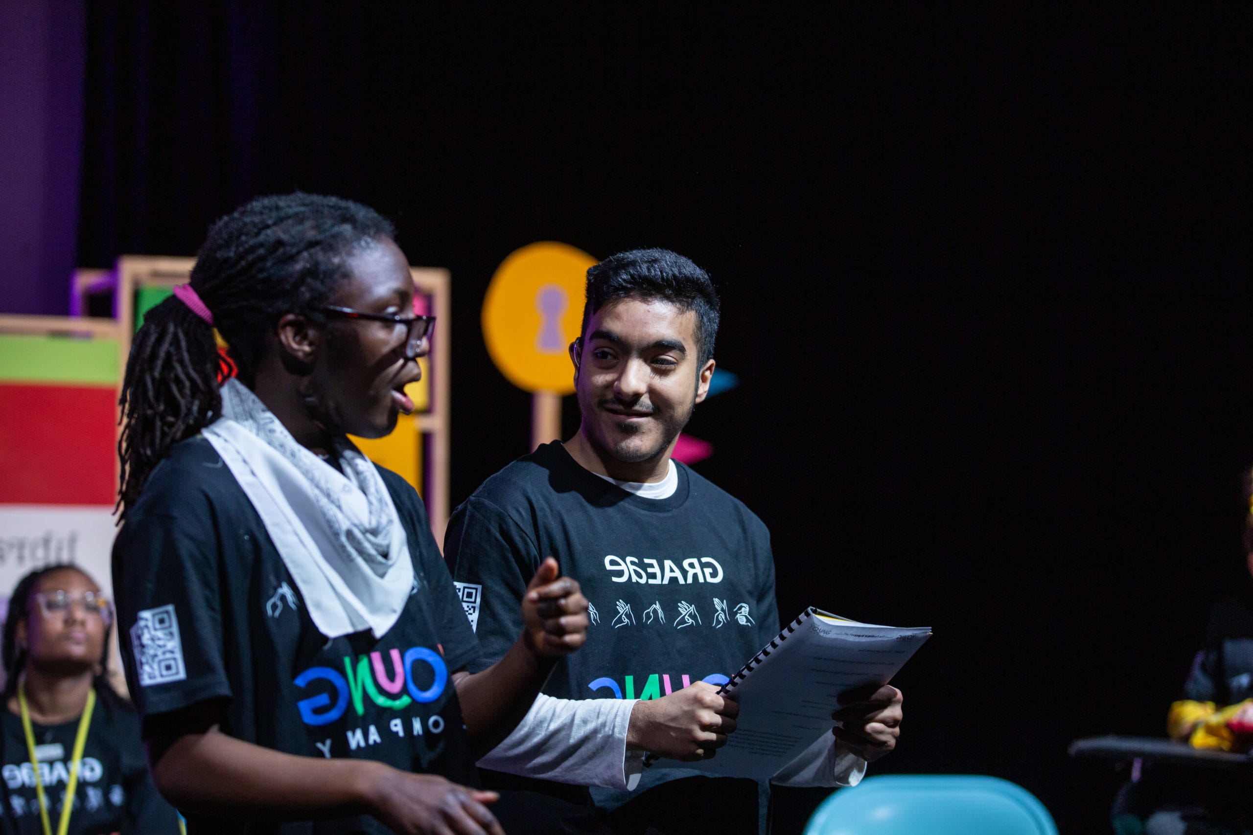 Two performers onstage. One is holding a script and smiling at the other, who appears to be smiling.