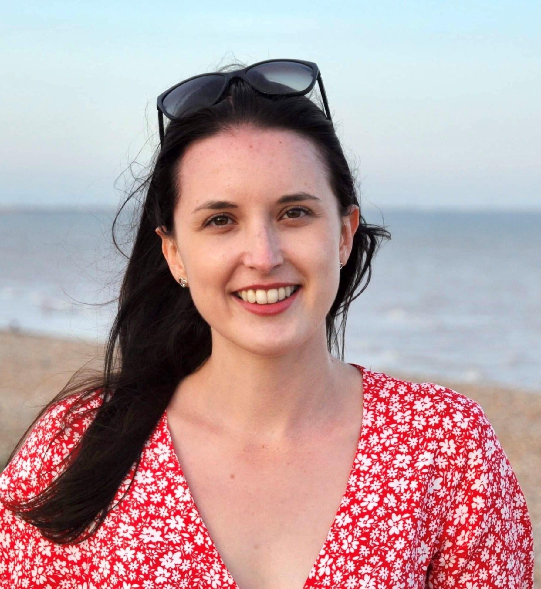 A white woman in her early thirties with long brown hair over her left shoulder, she wears a red flowery dress with sunglasses on her head