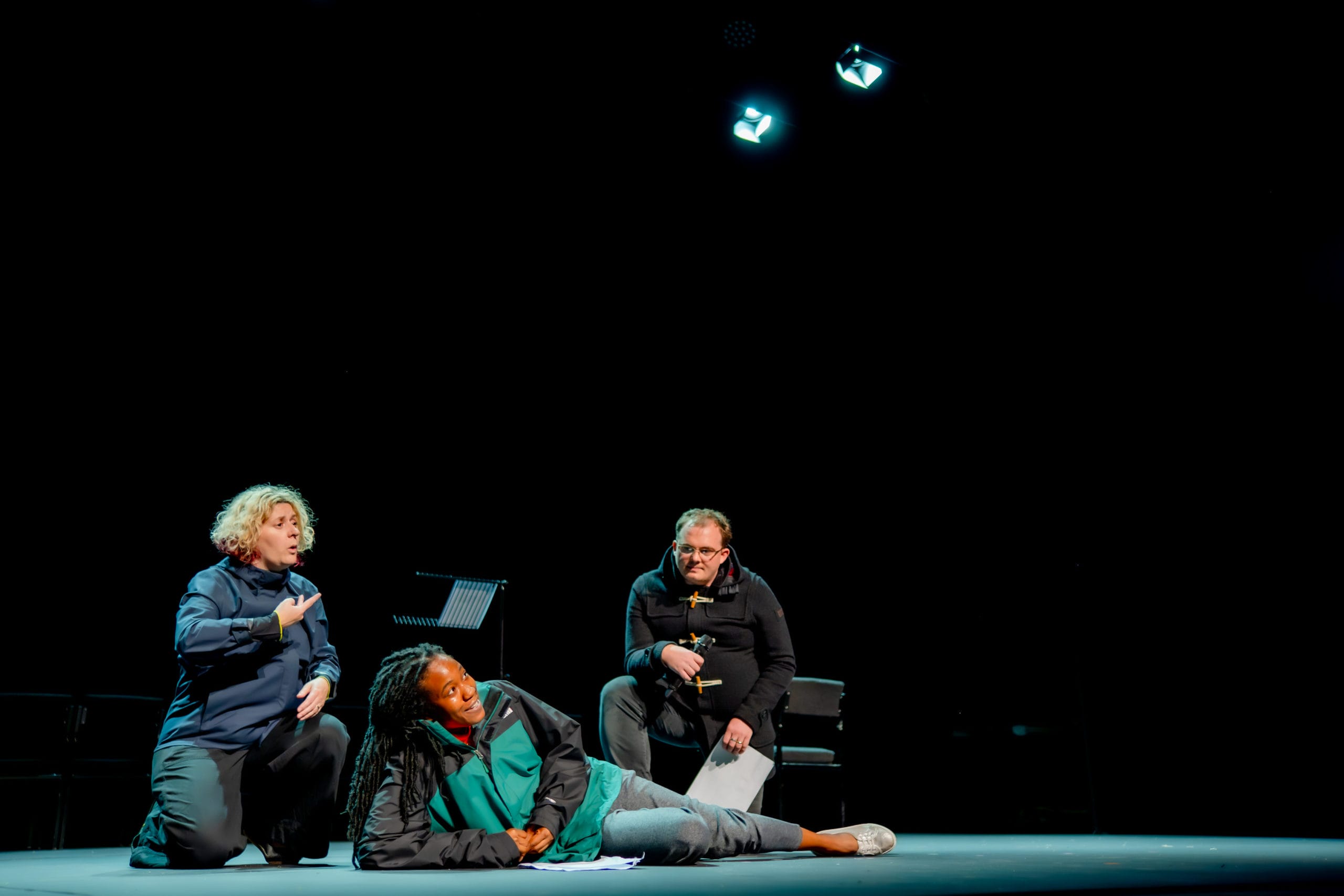 Three performers on the stage. Two of them are down on one knee, and the third is lying on the floor, propped up her elbow. One of the kneeling performers is signing.