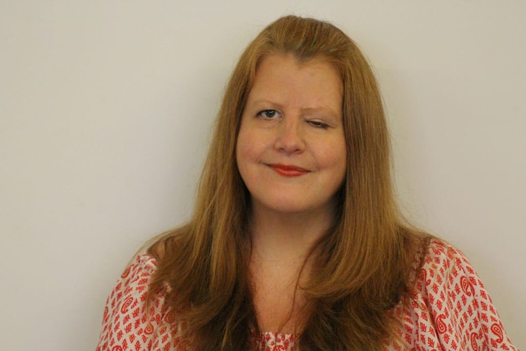 Jodi, a white woman with long blonde hair is stood in front of a plain white backdrop. She is smiling, wearing a white blouse with a red swirling pattern on it