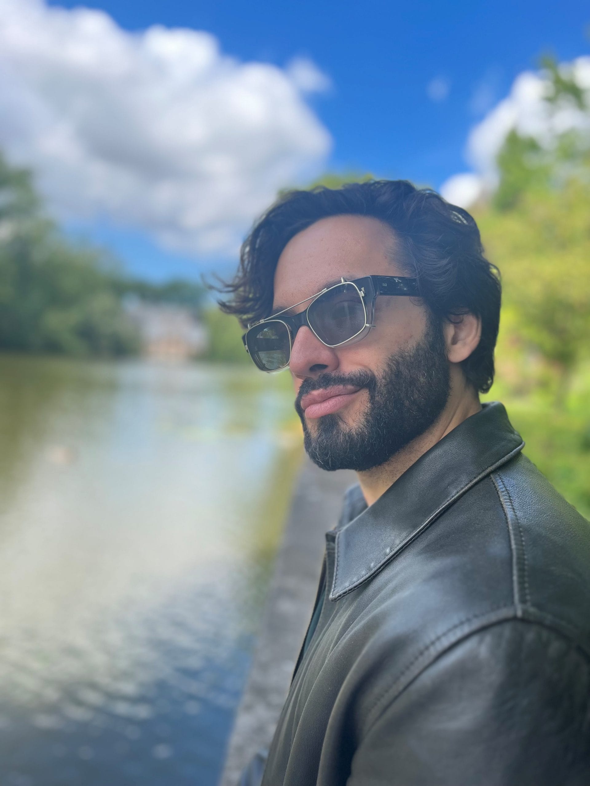 A man with dark brown hair and a beard wearing glasses with a leather jacket on. A river is in the background