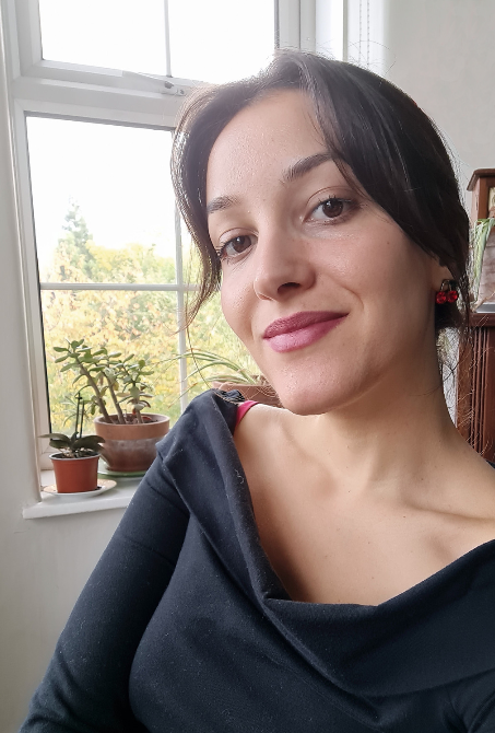 Selfie of a white woman in her early thirties smiling at the camera. She has brown straight hair in a ponytail and wears pink lipstick. There are pots with plants sitting on the windows in the background behind her.