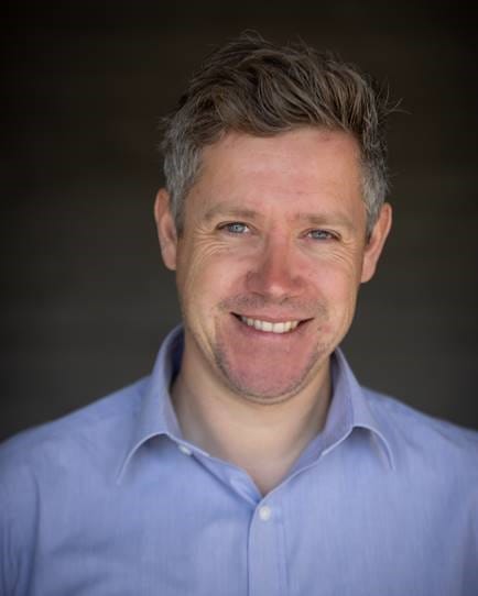 Matt, a white man with dark hair, is stood in front of a grey backdrop. He has a stubbly beard, and is smiling a broad smile. He is wearing a light blue work shirt, with the top button undone.