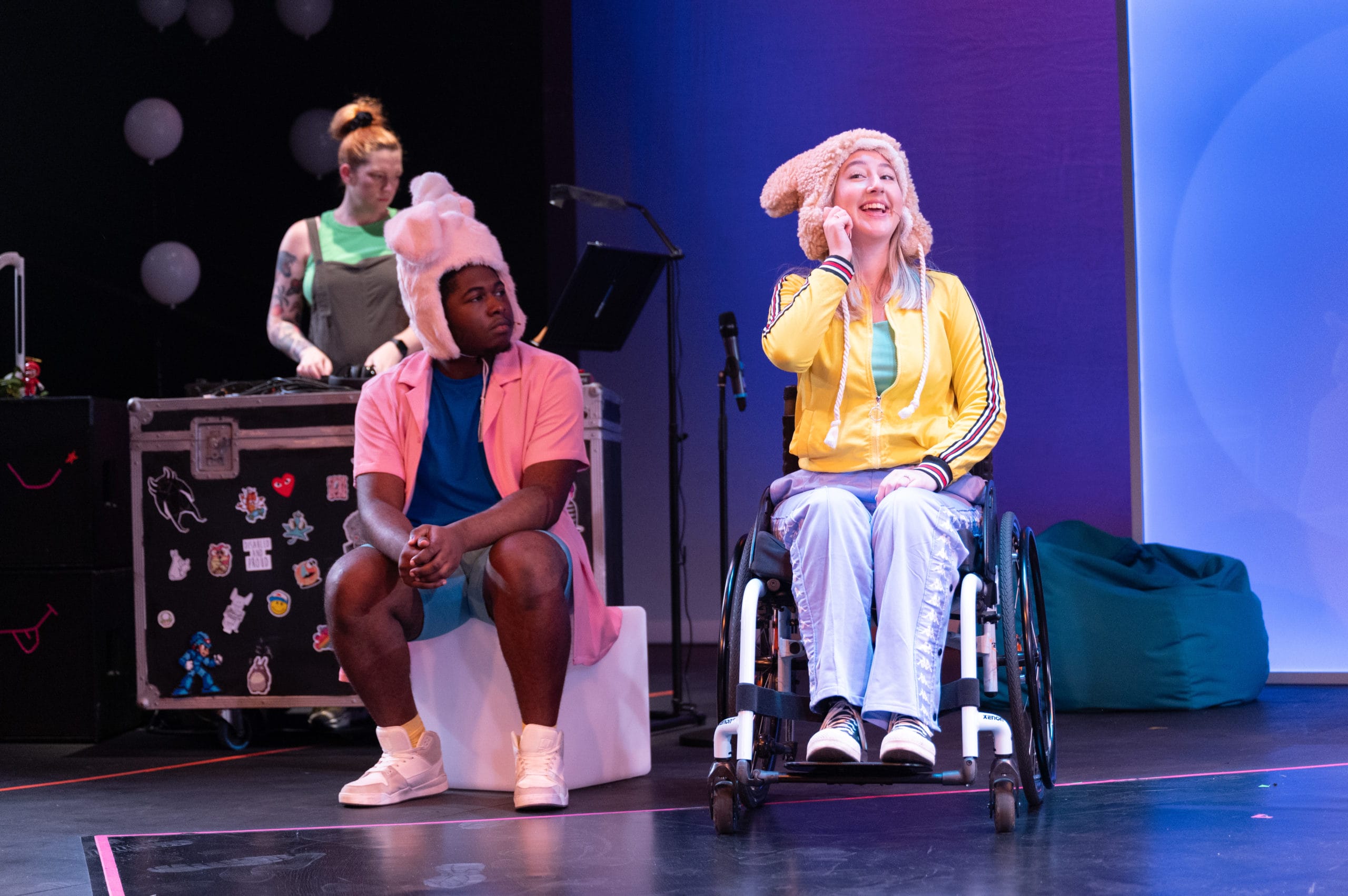Three performers on stage. The one in a wheelchair gestures to her ear, smiling. The second sits on a block watching her. The third is a DJ stood behind her decks.