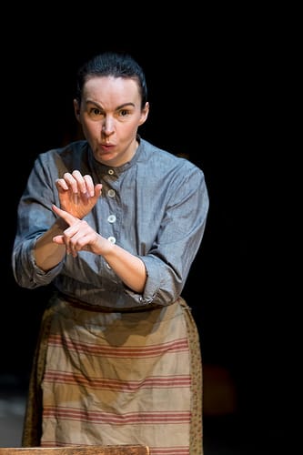 A female performer dressed in a grey shirt and floral skirt, with a striped apron tied around her waist. She is white and has dark brown hair pulled back in a bun.