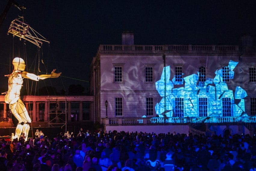 A huge puppet, glowing with golden light and operated by a crane, walks through a large crowd of people. There is a large building in the background that has blue light patterns projected onto it.