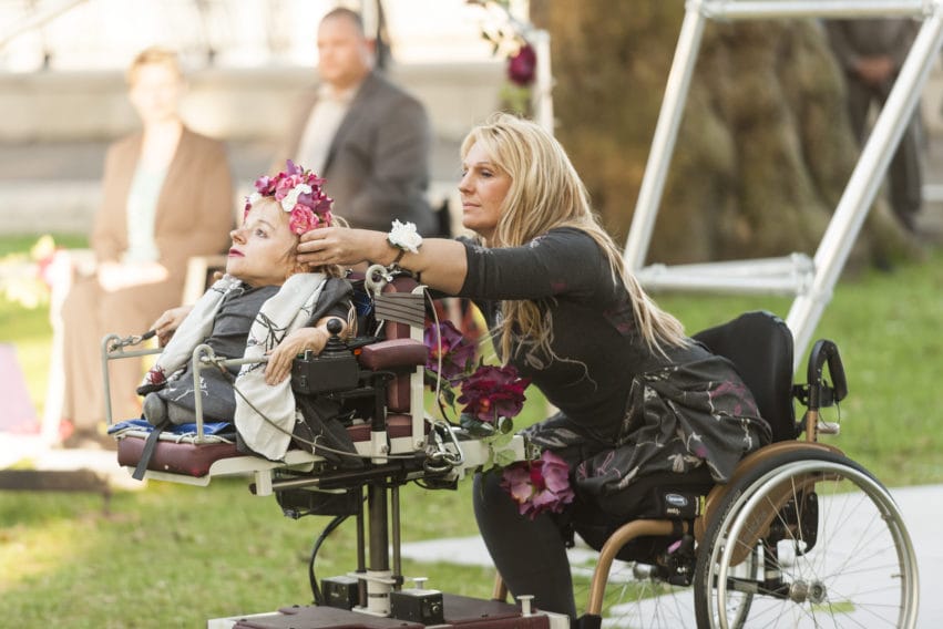 A female performer places a flower crown on the head of another.
