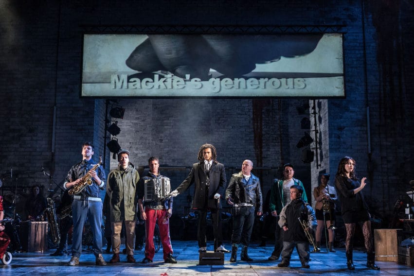 A group of performers stand on stage, facing out towards the audience. Some of them hold instruments.