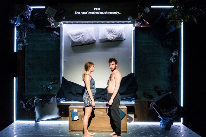 Two performers, a man and a woman, stand facing each other on stage. The man is looking out towards the audience and the woman is looking straight at him The stage is set behind them as a messy bedroom.