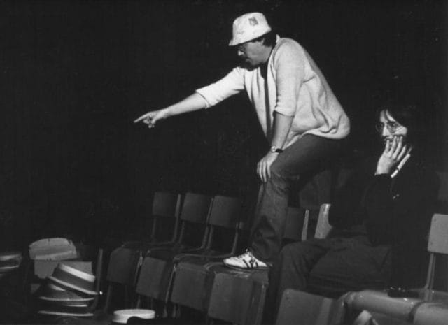 A black and white picture. Two men, one is sat on a row of chairs looking disinterested; the other is stood on the chairs pointing forward.